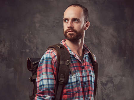 Portrait of a handsome tattooed traveler in a flannel shirt with a backpack, standing in a studio.の写真素材