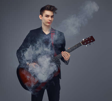 Handsome young musician with stylish hair in elegant clothes exhales smoke while playing acoustic...