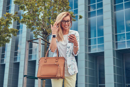 Fashionable female blogger in stylish clothes and glasses with a handbag, holds a smartphone,...