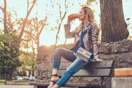 Trendy blonde female relaxes outdoor, drinking takeaway coffee while sitting on the bench in city park.の写真素材
