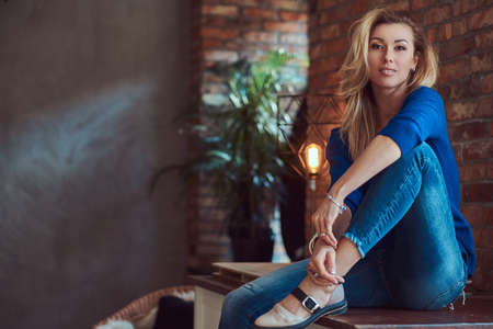 Beautiful blonde female sitting on a table against a brick wall in a studio with a loft interior.の写真素材