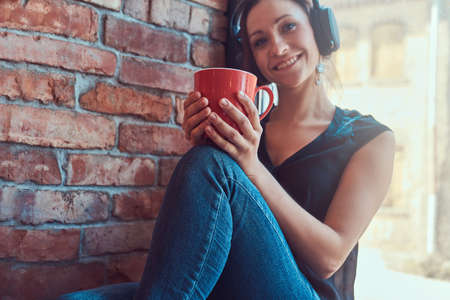Brunette female in black blouse listening music in headphones and holds a cup of coffee while...