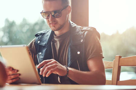 Handsome hipster with a stylish haircut and beard sits at a table in a roadside cafe, looks something in the tablet computer.の写真素材