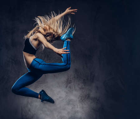 Young blonde ballerina in sportswear dances and jumps in a studio. Isolated on a dark background.