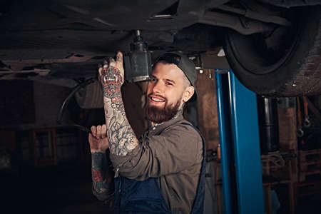 Auto mechanic in a uniform, repair the car with a screwdriver while standing under lifting car in the repair garage.の写真素材