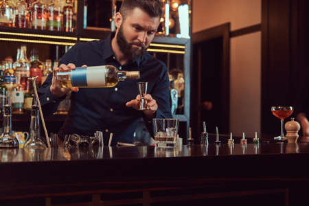 Stylish brutal barman in a black shirt makes a cocktail at bar counter background.