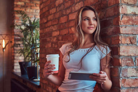 Portrait of a young blonde female holds a cup of a takeaway coffee and digital tablet while leaning against a brick wall in a room with loft interior.の写真素材