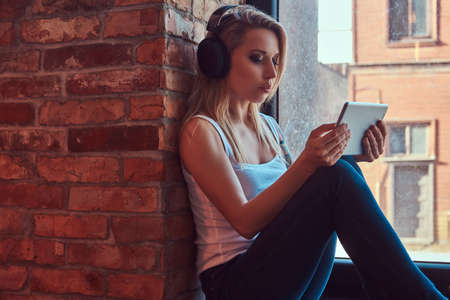 Portrait of a blonde tattoed hipster woman looking something on a digital tablet and listening music while sitting on a window sill in a room with loft interior.の写真素材