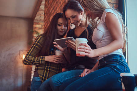 Group of female friends in casual clothes discussing while looking something on a digital tablet in a room with loft interior.の写真素材