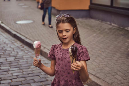 Beauty little girl in a fashionable dress holds two ice cream cones, standing on the summer city street.の写真素材