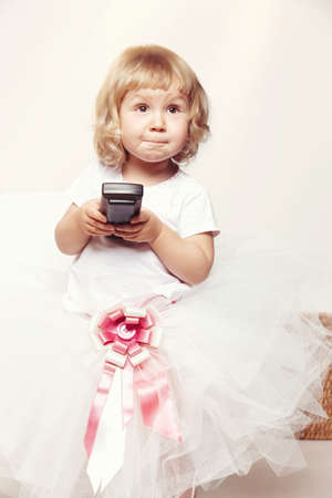 Portrait of a cute little girl in a white dress holds a tv remote on white background.
