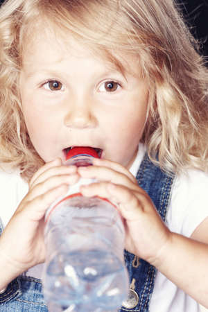 Portrait of a cute little girl in denim overalls, drink water while sitting in studio.の写真素材
