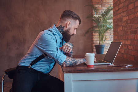 Handsome tattooed hipster in a shirt and suspenders sitting at the desk, working on a laptop, holds...