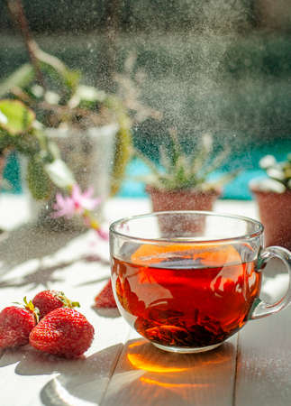 Close-up image of glass cup of tea, red strawberries, flowers in pots on a light wooden table.の写真素材