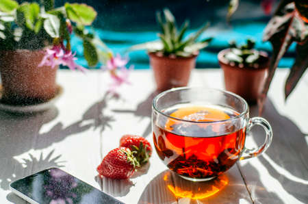 Close-up image of glass cup of tea, red strawberries, smartphone and flowers in pots on a light wooden table.の写真素材
