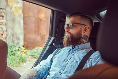 Old-fashioned tattooed hipster guy in a shirt with suspenders sitting in a luxury car on back seat.