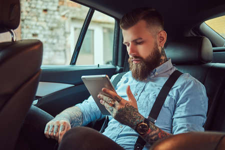 Old-fashioned tattooed hipster guy in a shirt with suspenders, using a tablet while sitting in a...