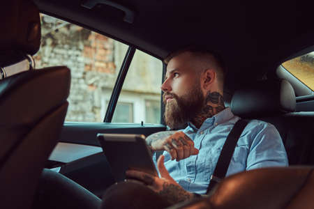 Old-fashioned tattooed hipster guy in a shirt with suspenders, holding a tablet while sitting in a...