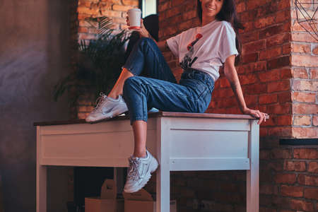 Cropped image of a cool modern girl wearing a white top and jeans sitting on a table with a cup of takeaway coffee in studio with loft interior.の写真素材