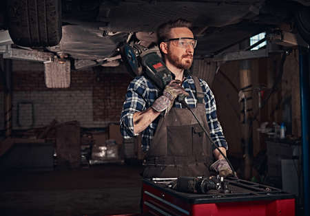 Mechanic in a uniform and safety glasses holds an angle grinder while standing under lifting car in...