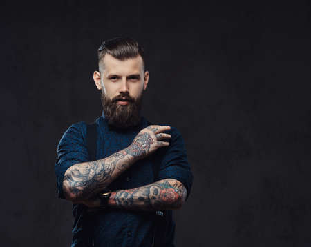 Handsome old-fashioned hipster in a blue shirt and suspenders, standing with crossed arms in a studio.の写真素材