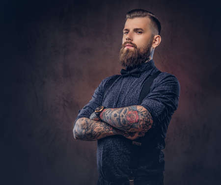 Portrait of a handsome old-fashioned hipster in a blue shirt and suspenders, standing with crossed arms in a studio.の写真素材