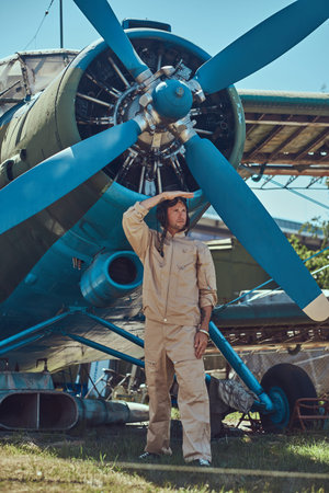Valiant handsome pilot in a full flight gear standing near a retro military airplane and looks away.の写真素材