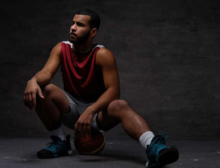 Young African-American basketball player in sportswear sitting on a ball on a dark background.の写真素材