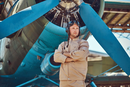 Valiant handsome pilot in a full flight gear standing with crossed arms near military airplane.