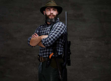 Portrait of a bearded hunter in a fleece shirt and hat standing with a rifle behind his back.の写真素材