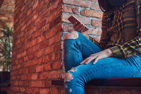Young brunette student girl in casual clothes listening music while using a smartphone while sitting on a window sill in a room with loft interior.の写真素材