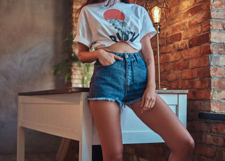Cropped image of a sexy slim cool girl in a white top and denim shorts, leaning on a table in studio with loft interior.の写真素材