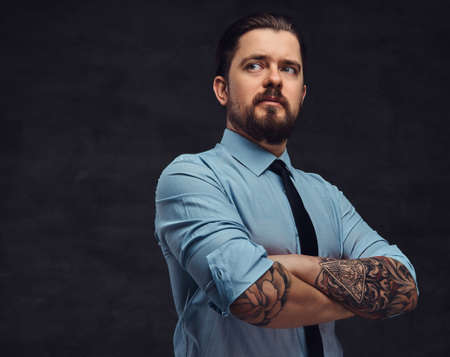 Portrait of a tattooed handsome middle-aged man with beard and hairstyle dressed in a blue shirt and tie, pose in a studio with crossed arms.の写真素材