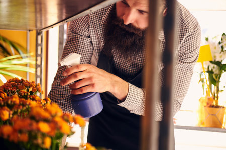 Professional male florist with beard and tattoo on his hand wearing uniform taking care of flowers...