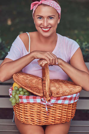 Happy beautiful redhead female wearing casual clothes holds a picnic basket while sitting on a bench in a park.の写真素材