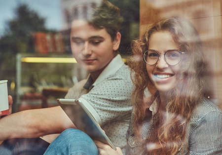 Couple of young students drinking coffee and using a digital tablet while sitting on a window sill...
