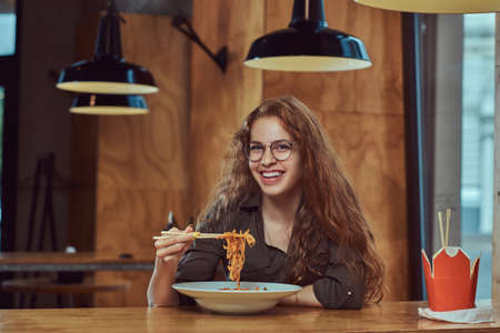 Young redhead female eating spicy noodles in an Asian restaurant.