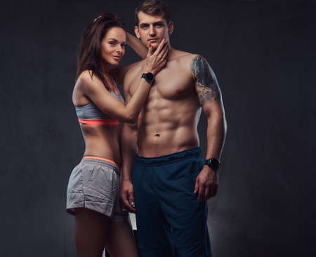 Attractive couple, a slim brunette female wearing sports bra and shorts and handsome shirtless guy cuddling in a studio on a dark textured background.の写真素材