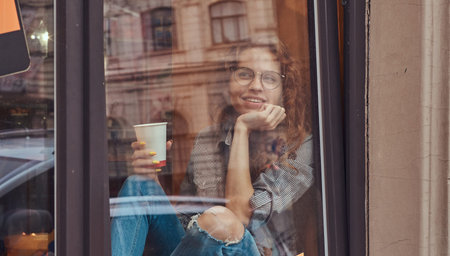 Young redhead curly girl wearing casual clothes and glasses sitting on a window sill with a takeaway...