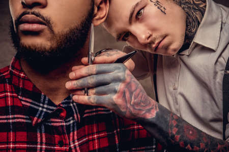 Handsome bearded African American guy in a fleece shirt and cap getting a haircut by an old-fashioned professional hairdresser does haircut.の写真素材