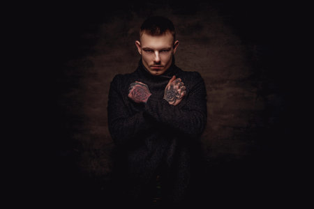 Tattoed young guy in a black jacket posing with crossed arms at the studio. Isolated on dark textured background.の写真素材