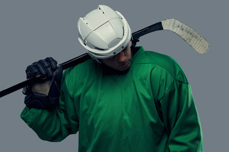 Hockey player wearing green protective gear and white helmet standing with the hockey stick. Isolated on a gray background.の写真素材