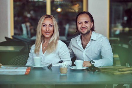 Happy attractive couple - charming blonde female dressed in a white blouse and bearded male with a stylish haircut dressed in a white shirt during a date at a cafe outdoors.の写真素材