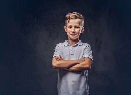 Cute little boy dressed in a white t-shirt standing with crossed arms in a studio. Isolated on dark textured background.の写真素材