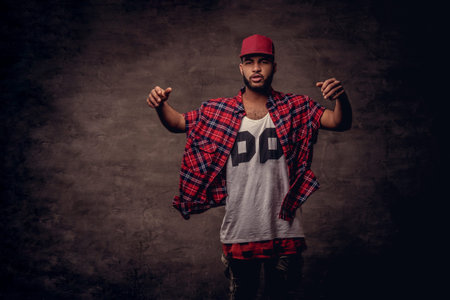 Portrait of an African-American dancer guy dressed in a red fleece shirt and cap at the studio. Isolated on dark textured background.の写真素材