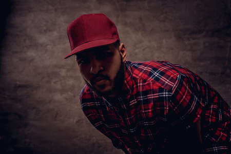 Portrait of an African-American hipster guy dressed in a red fleece shirt and cap at the studio. Isolated on dark textured background.の写真素材
