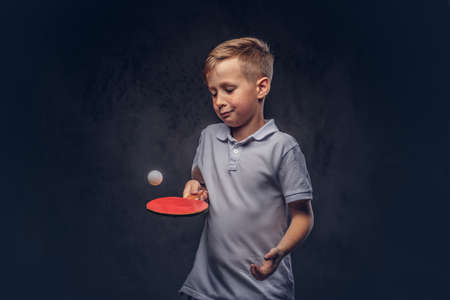 Cute redhead boy dressed in a white t-shirt playing table tennis in a studio. Isolated on dark textured background.の写真素材