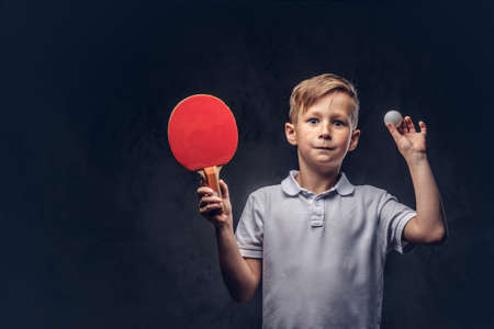 Cute redhead boy dressed in a white t-shirt holds a table tennis racket and ball in a studio. Isolated on a dark textured background.の写真素材