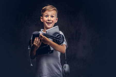 Joyful Cute schoolboy dressed in a white t-shirt, holds full digital set for entertainment at a studio. Isolated on dark textured background.の写真素材