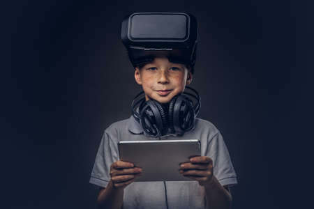 Portrait of a cute little boy dressed in a white t-shirt, with virtual reality glasses and headphones is holding a digital tablet at a studio, isolated on dark background. Education and technology concept.の写真素材